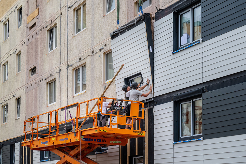 Zwei Bauarbeiter stehen auf einer Hebebühne und bringen vorgefertigte Fassadenmodule an.